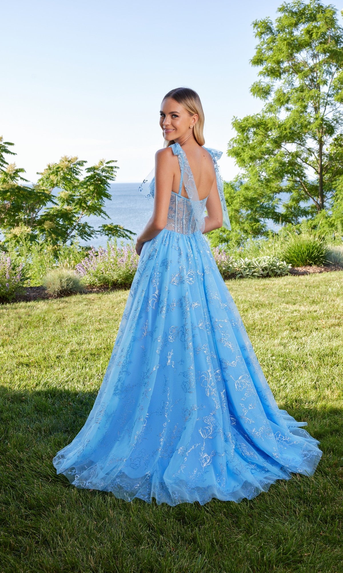 A woman stands on grass near water, wearing the Morilee 49185 long prom dress—a light-blue gown with silver patterns, an a-line tulle skirt, and shoulder bows. She smiles over her shoulder, with trees and shrubs behind her.